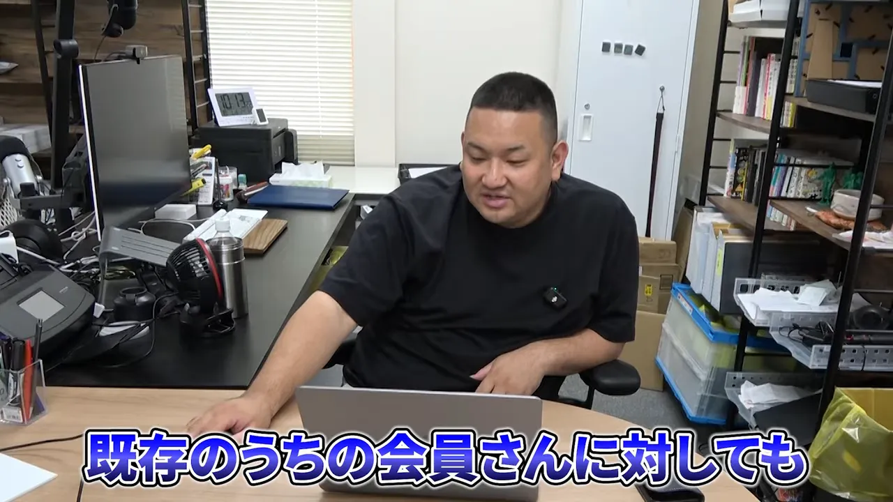 オフィスのテーブルでノートPCを見ながら説明する様子(青い字幕あり)