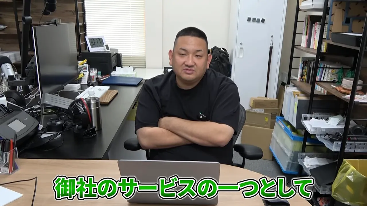オフィスの机で正面を向いて説明する男性(字幕:「御社のサービスの一つとして」)