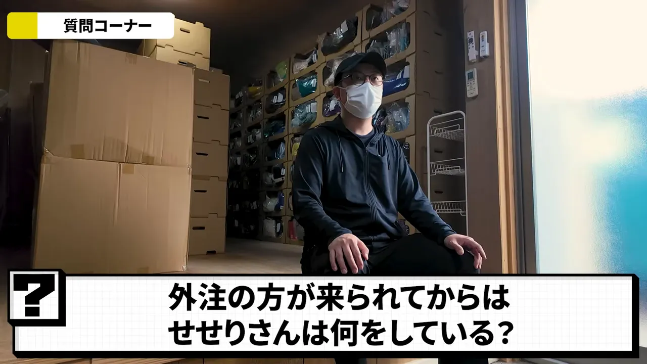 在庫箱と棚が並ぶ倉庫で座り説明する人物。外注と分業する運営のイメージ