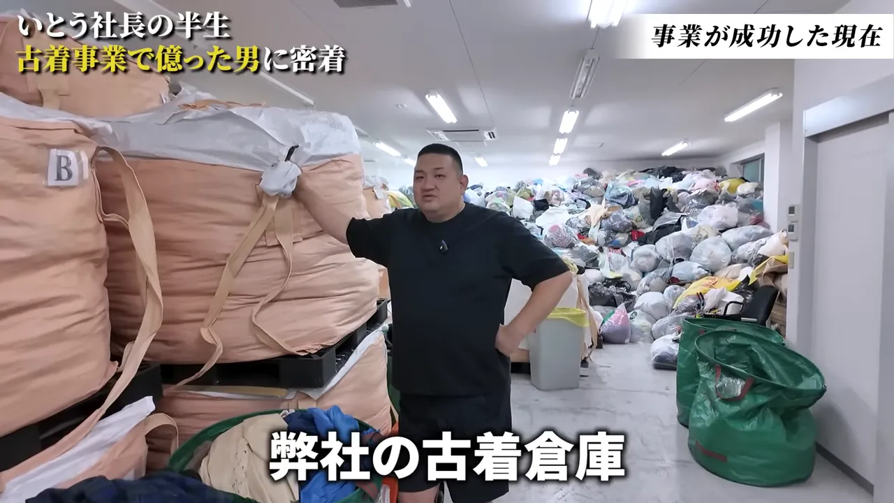 倉庫内で腕を置いて立つ代表と、奥に積まれた大量の古着袋が見える全景写真