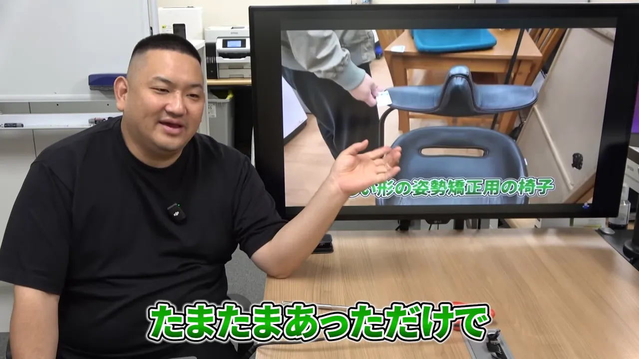 モニターに姿勢矯正用の珍しい形の椅子が映り、解説者が身振りで説明している様子