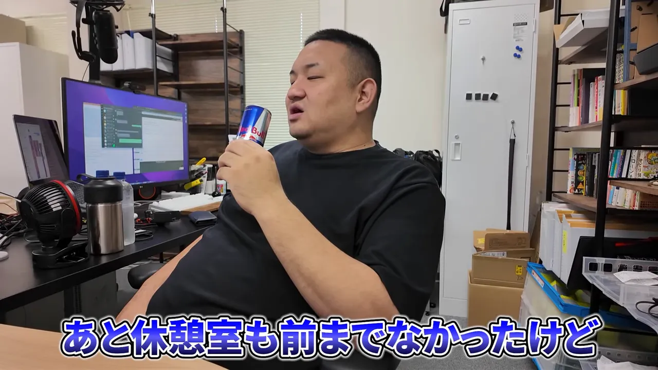 デスク越しに缶飲料を手に話す社長と、奥の棚や箱が見えるオフィス風景